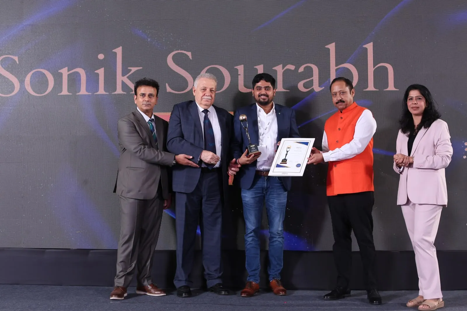 Sonik Sourabh holding a trophy and certificate on stage, flanked by summit hosts